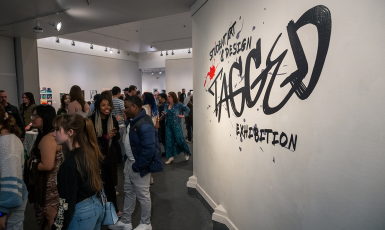 Art exhibit room filled with people. Paint on wall reads, "Student Art & Design TAGGED Exhibition"