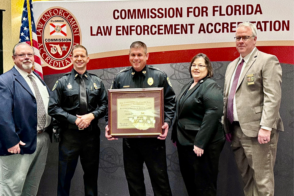 UWF Police Chief holds framed accreditation award with two people on his left and right. Text on wall behind them reads, "Commission for Florida Law Enforcement Accreditation."