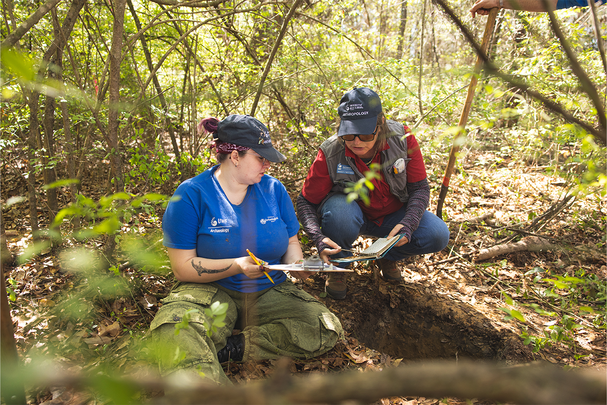 Two people sit on the ground in the woods writing on a clipboard during an archaeology dig.