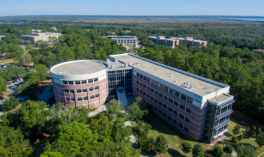 Aerial shot of UWF's Hal Marcus College of Science and Engineering building in the shape of a zero and one, surrounded by greenery and a blue sky.