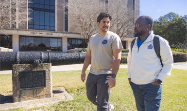 Two UWF military students in civilian clothing walk on the Cannon Green in front of Annin's Cannon and the Pace Library.