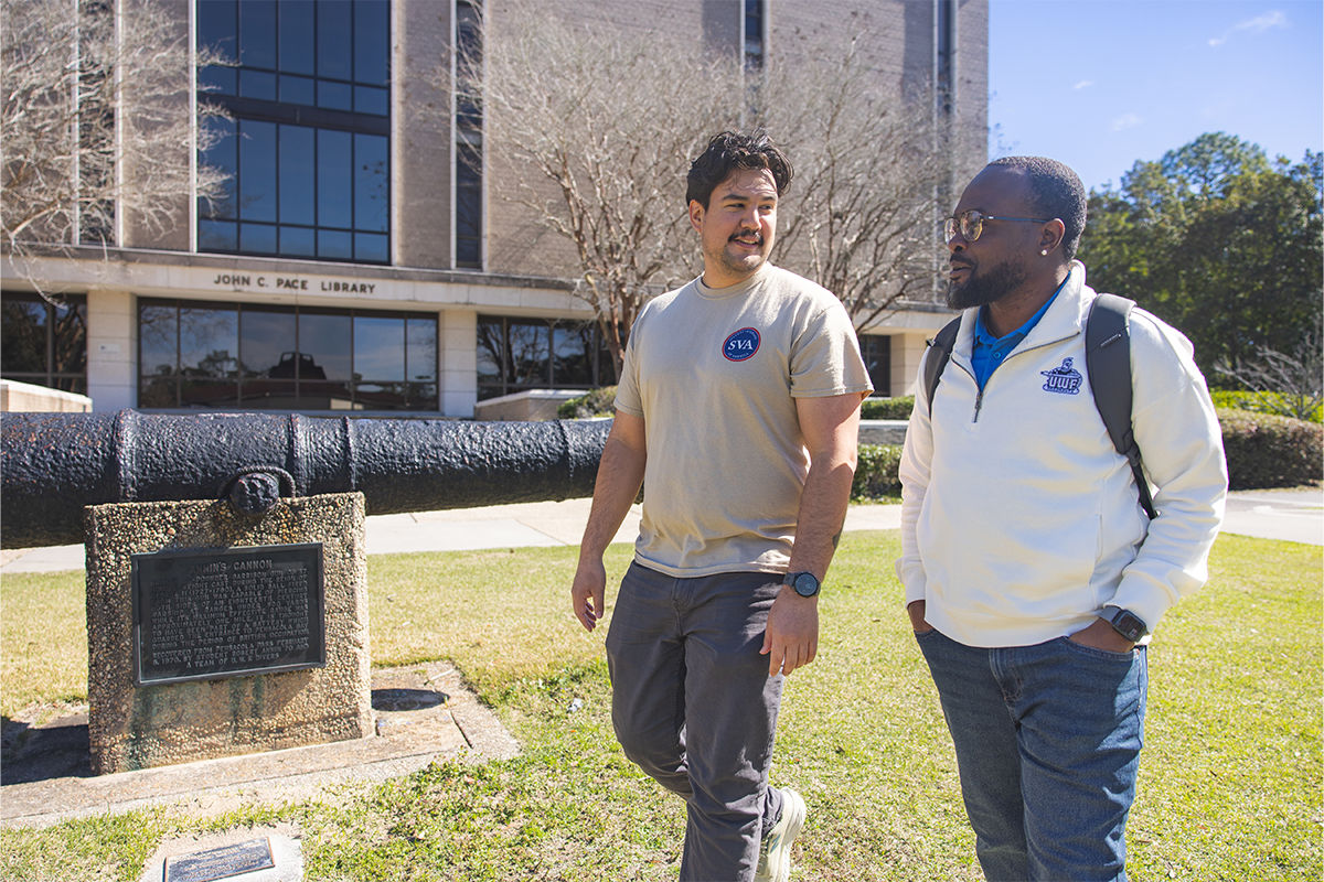 Two UWF military students in civilian clothing walk on the Cannon Green in front of Annin's Cannon and the Pace Library.