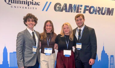 Four students in business attire stand together smiling in front of a backdrop that says, "Quinnipiac University Game Forum."
