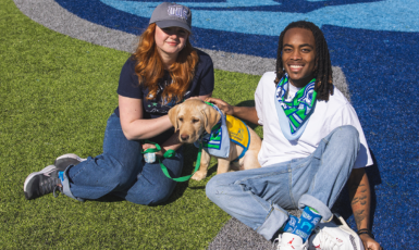 Two students and a puppy in UWF Day of Giving merch sitting on a football field with the bottom half of the Argonaut helmet logo visible on the field behind them.