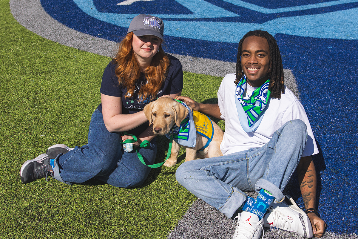 Two students and a puppy in UWF Day of Giving merch sitting on a football field with the bottom half of the Argonaut helmet logo visible on the field behind them.
