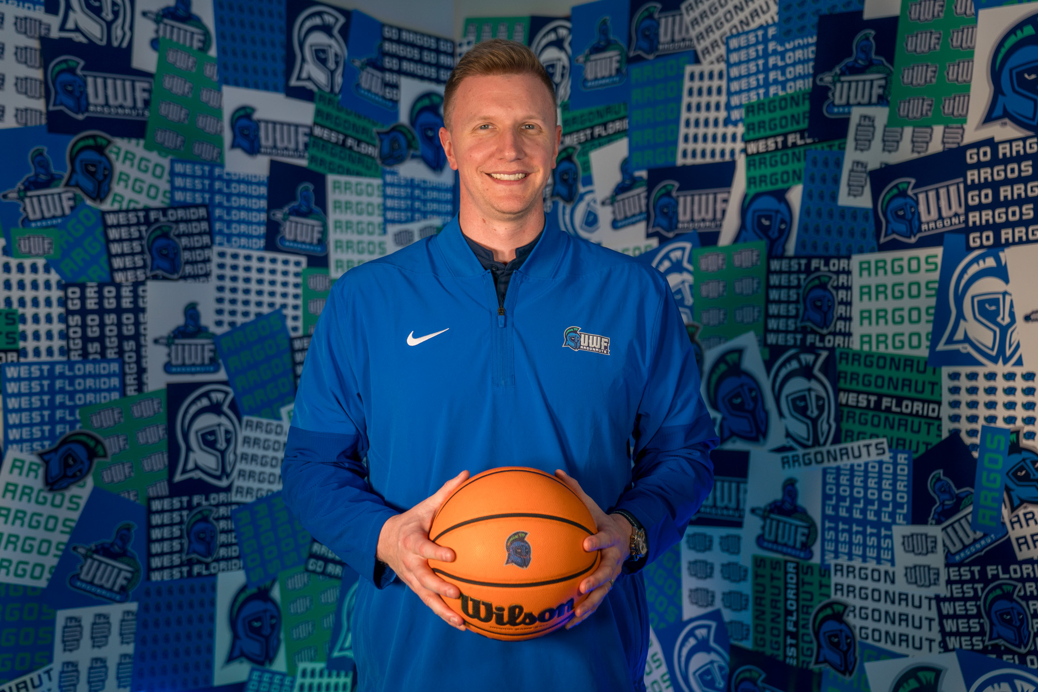 Basketball coach holds basketball in hands in front of a collage wall with Argo graphics.