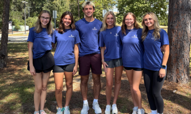 Six students stand together in matching blue t-shirts, smiling, with trees behind them.
