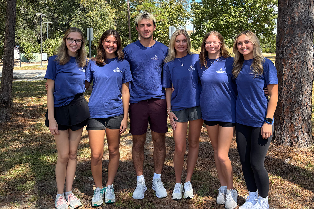 Six students stand together in matching blue t-shirts, smiling, with trees behind them.