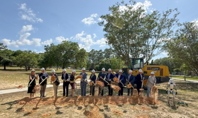 Photo: Thirteen people in a line with color-white hard hats scoop up dirt with shovels in front of a piece of construction machinery and next to a humanoid robot.
