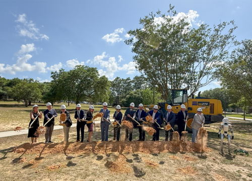 Photo: Thirteen people in a line with color-white hard hats scoop up dirt with shovels in front of a piece of construction machinery and next to a humanoid robot.
