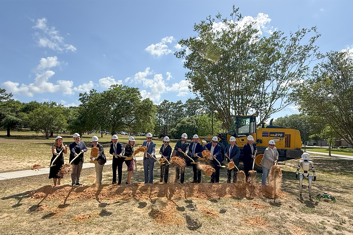 Photo: Thirteen people in a line with color-white hard hats scoop up dirt with shovels in front of a piece of construction machinery and next to a humanoid robot.