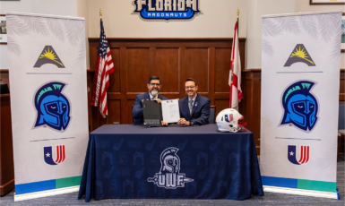 Two people sit behind a table with a UWF dark blue tablecloth and West Florida Argonauts sign above their heads. Two posters on either side have the ASUN, UWF and UAC logos.