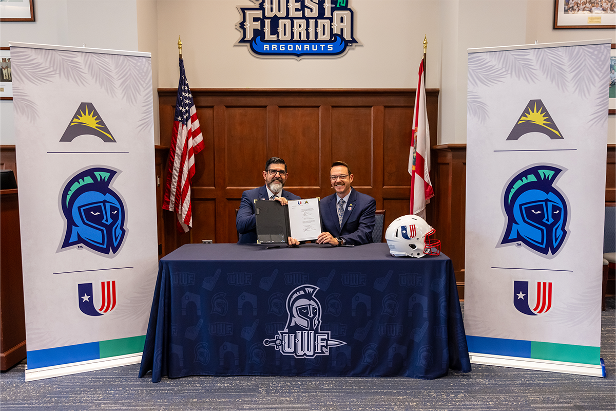 Two people sit behind a table with a UWF dark blue tablecloth and West Florida Argonauts sign above their heads. Two posters on either side have the ASUN, UWF and UAC logos.