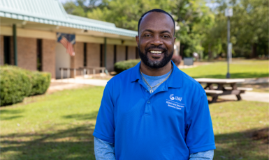 Photo: Person in color-blue shirt with logo text - UWF Military and Veterans Resource Center poses in front of a building, smiling.