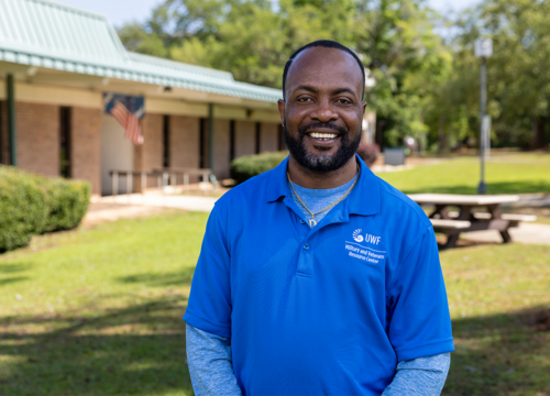 Photo: Person in color-blue shirt with logo text - UWF Military and Veterans Resource Center poses in front of a building, smiling.