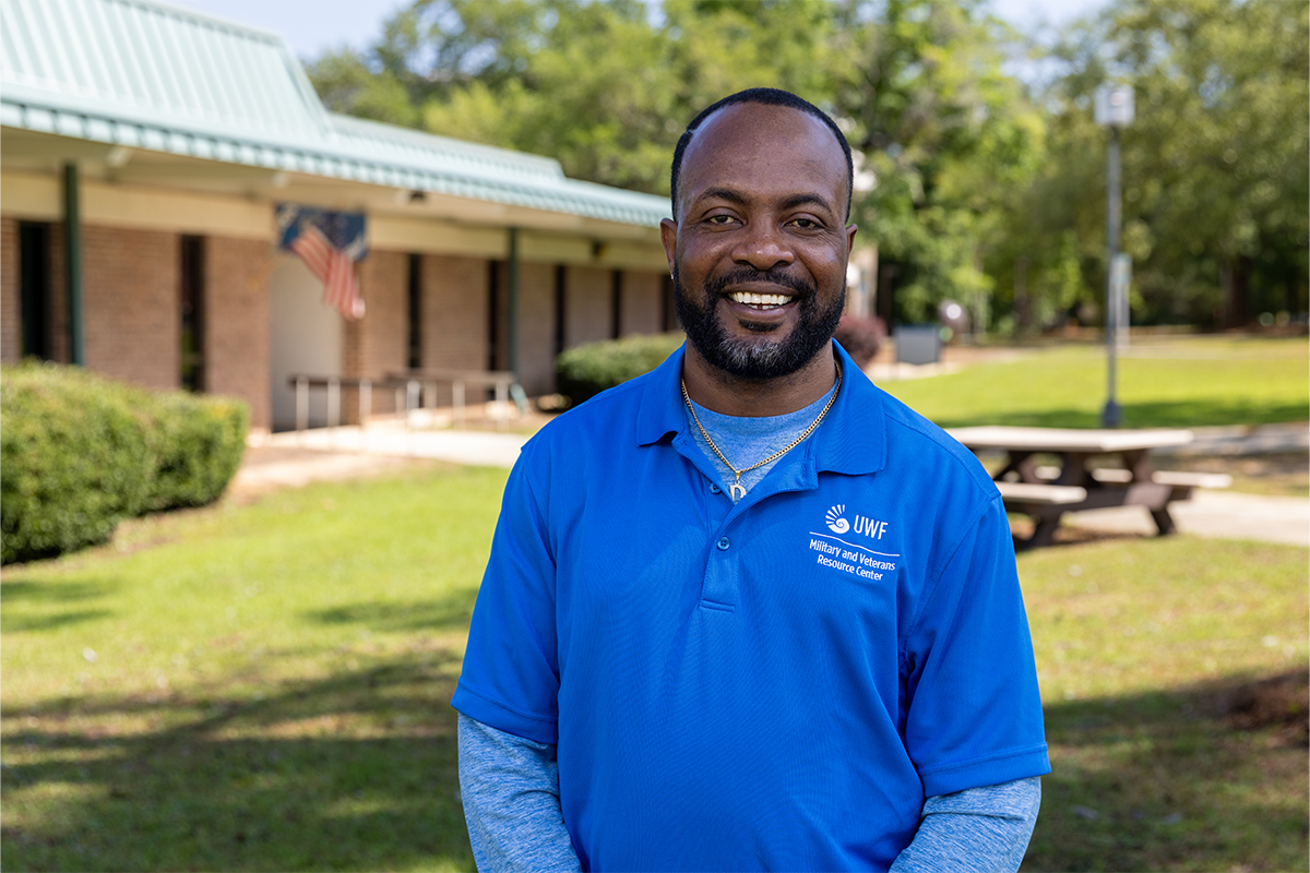 Photo: Person in color-blue shirt with logo text - UWF Military and Veterans Resource Center poses in front of a building, smiling.