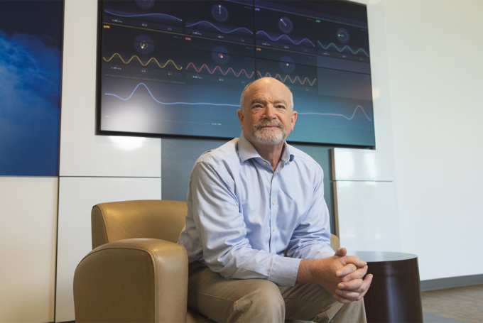 Dr. James Arruda sits in a leather chair in front of a large TV screen with EEG activity displayed.