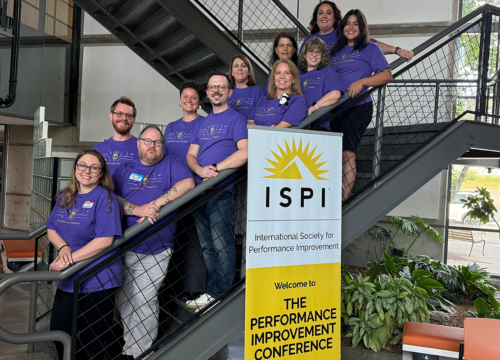 Two professors and nine students pose on a staircase behind a banner that reads: "ISPI International Society for Performance Improvement. Welcome to The Performance Improvement Conference."
