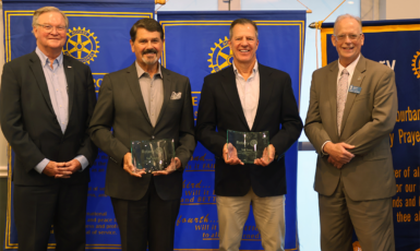 Four people stand in suits in front of color-blue banners. The middle two hold glass awards.