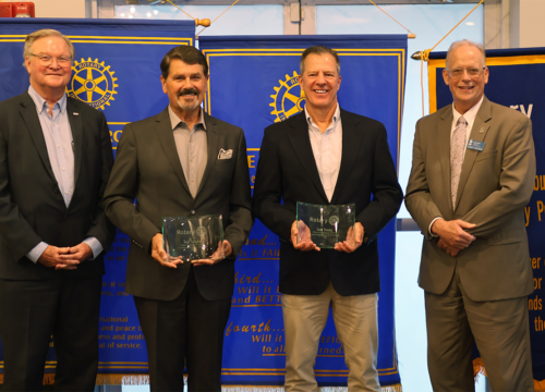 Four people stand in suits in front of color-blue banners. The middle two hold glass awards.