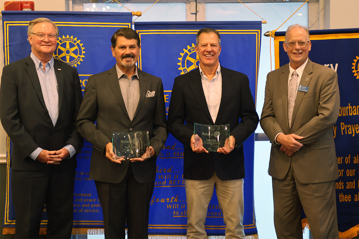Four people stand in suits in front of color-blue banners. The middle two hold glass awards.