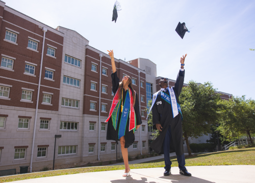 Two graduates throw their graduation caps in the air in front of a dormitory.