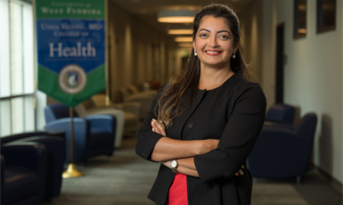 Dr. Karishma Chhabria Unrue stands smiling with arms crossed inside a lounge with a banner in the background with text: "University of West Florida Usha Kundu, MD College of Health"