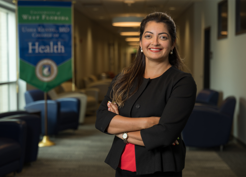 Dr. Karishma Chhabria Unrue stands smiling with arms crossed inside a lounge with a banner in the background with text: "University of West Florida Usha Kundu, MD College of Health"