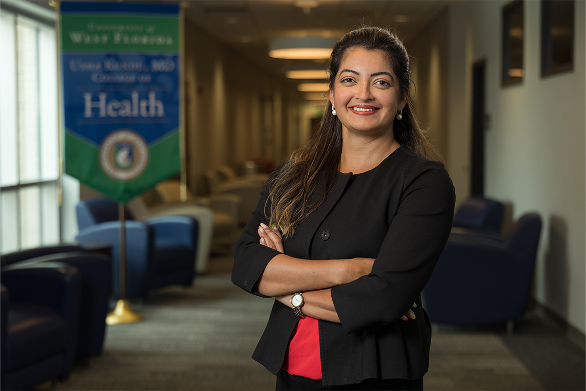 Dr. Karishma Chhabria Unrue stands smiling with arms crossed inside a lounge with a banner in the background with text: "University of West Florida Usha Kundu, MD College of Health"