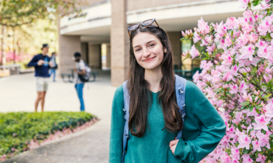 UWF student stands smiling with one arm on hip in front of the library and a bush with pink flowers