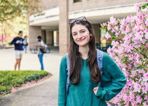 UWF student stands smiling with one arm on hip in front of the library and a bush with pink flowers