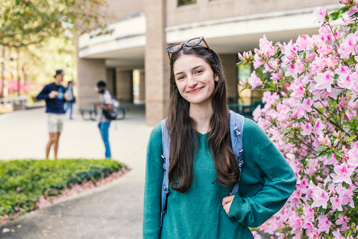 UWF student stands smiling with one arm on hip in front of the library and a bush with pink flowers