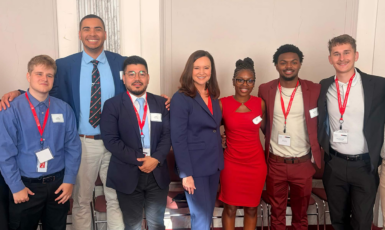 Six students in professional attire pose on either side of Senator Ashley Moody.