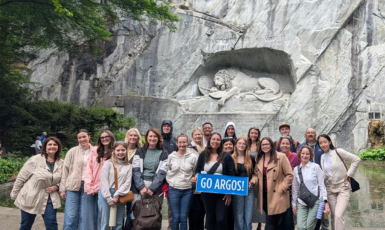 Group of students and faculty pose in front of a stone wall with a lion carved into it. Students hold a banner that says, "GO ARGOS!"