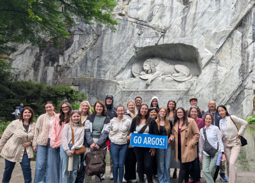 Group of students and faculty pose in front of a stone wall with a lion carved into it. Students hold a banner that says, "GO ARGOS!"