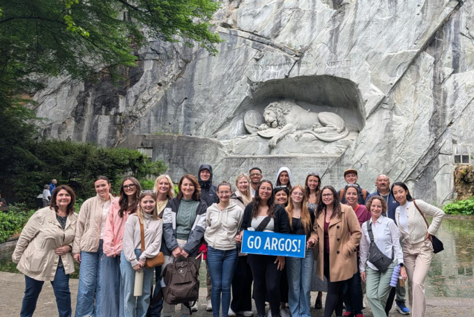 Group of students and faculty pose in front of a stone wall with a lion carved into it. Students hold a banner that says, "GO ARGOS!"