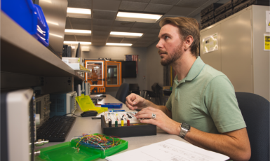 A student works at an electronics lab workstation assembling and testing a circuit board while monitoring equipment and a computer display.