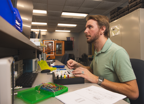 A student works at an electronics lab workstation assembling and testing a circuit board while monitoring equipment and a computer display.