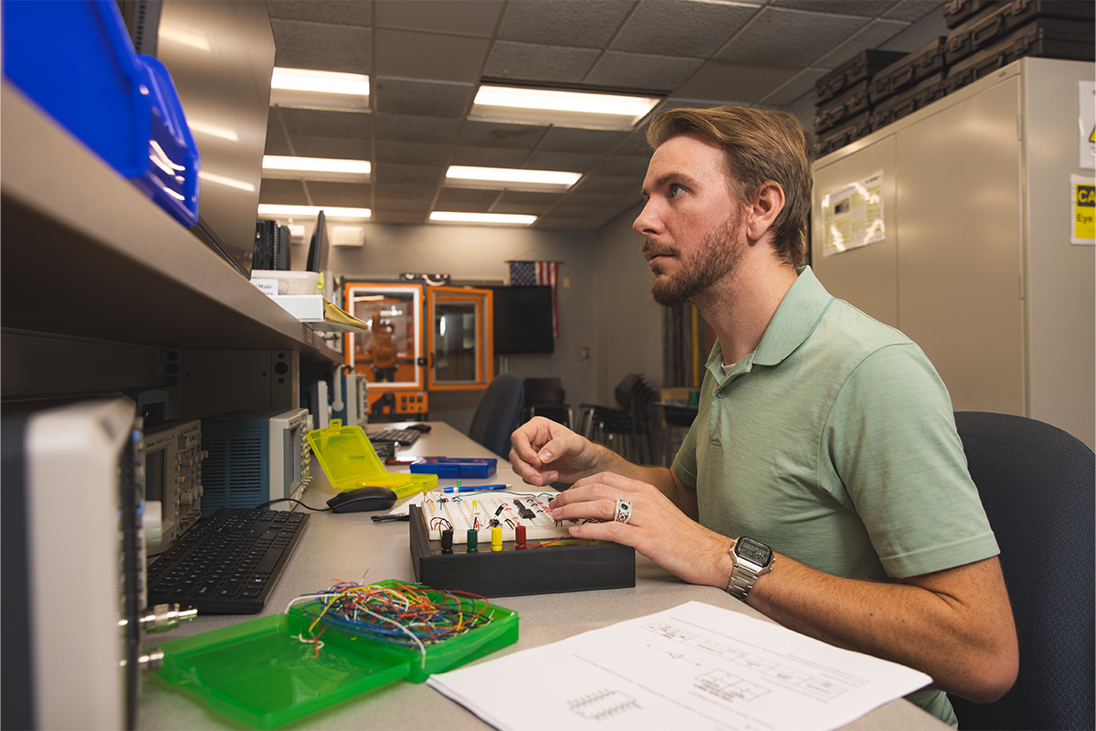 A student works at an electronics lab workstation assembling and testing a circuit board while monitoring equipment and a computer display.