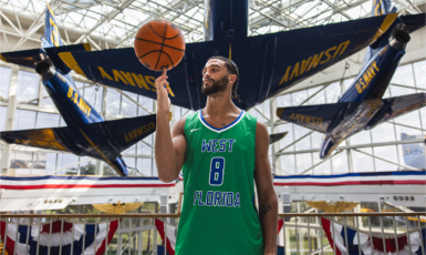UWF men's basketball player with jersey number 8 spins a basketball on his finger while standing in front of a U.S. Navy Blue Angels aircraft.
