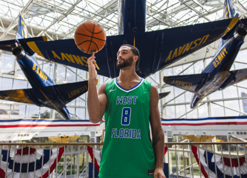 UWF men's basketball player with jersey number 8 spins a basketball on his finger while standing in front of a U.S. Navy Blue Angels aircraft.
