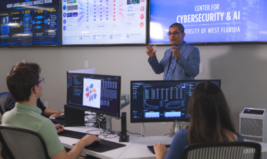 Professor teaches a cybersecurity class in front of students at desks with computers and screens on the wall behind him.