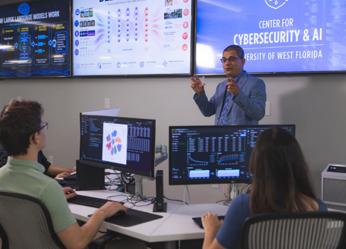 Professor teaches a cybersecurity class in front of students at desks with computers and screens on the wall behind him.