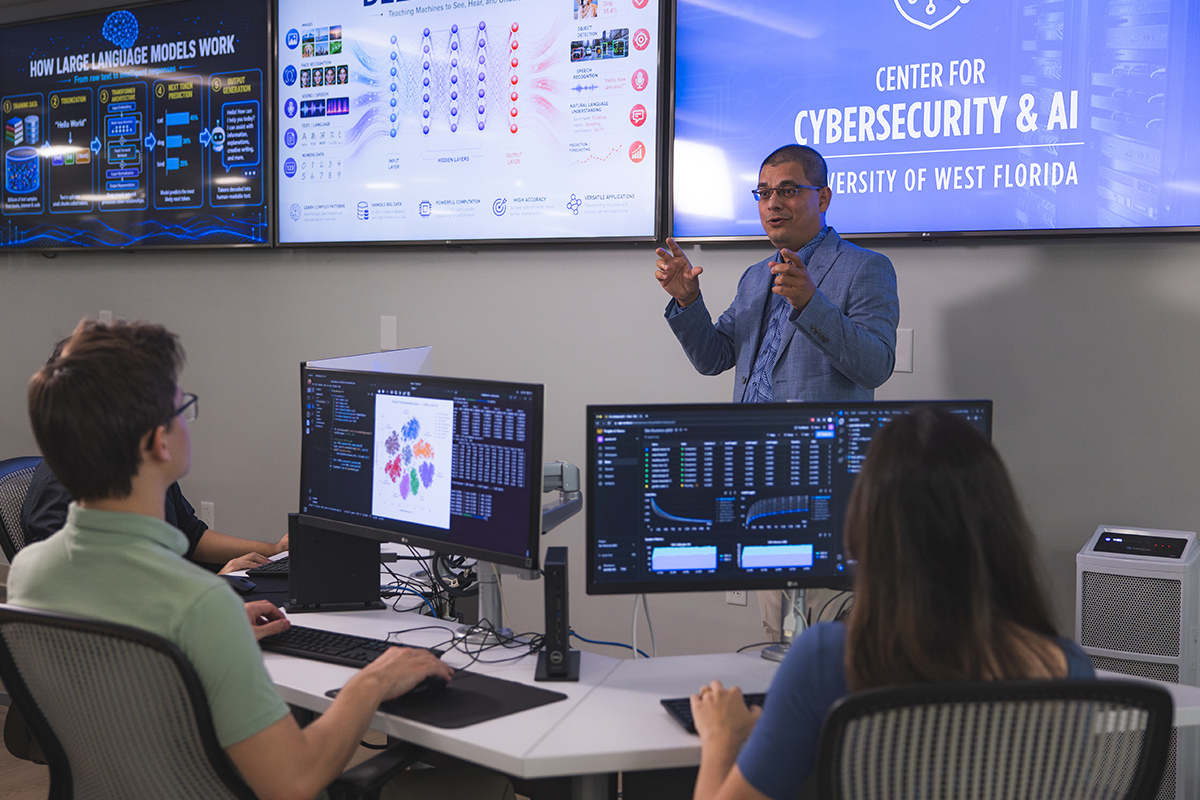 Professor teaches a cybersecurity class in front of students at desks with computers and screens on the wall behind him.