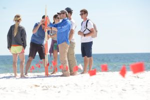 Professor leading an outdoor lesson with 5 students at the beach on how to use ranging poles while mapping.