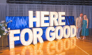 Giant display letters spell out HERE FOR GOOD. A bouquet of blue flowers stand to the left while two people pose on the right.