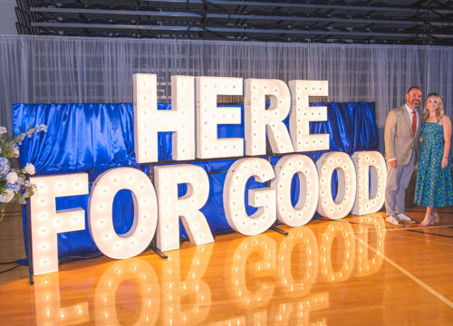 Giant display letters spell out HERE FOR GOOD. A bouquet of blue flowers stand to the left while two people pose on the right.