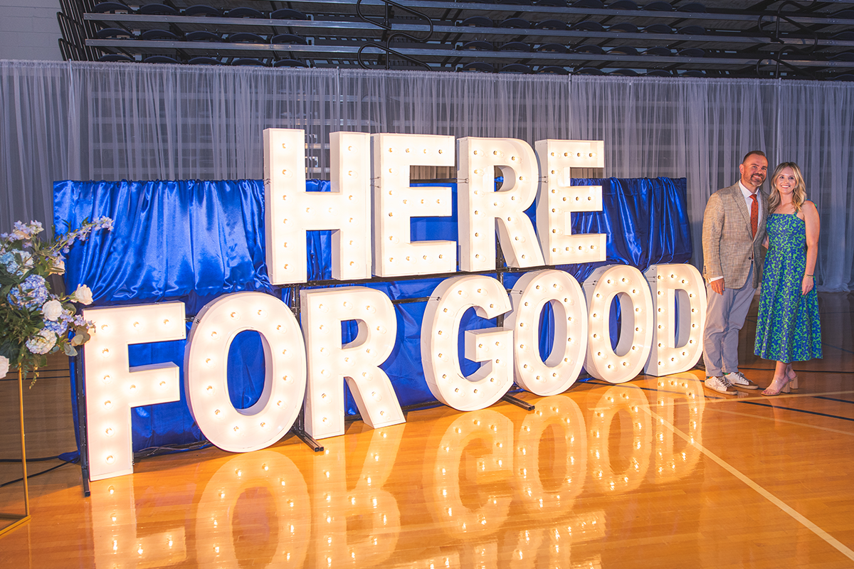 Giant display letters spell out HERE FOR GOOD. A bouquet of blue flowers stand to the left while two people pose on the right.