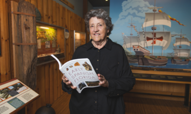 Dr. Judy Bense holds her book, "Early Spanish Florida," inside a museum with a Spanish ship painted on the wall behind her.
