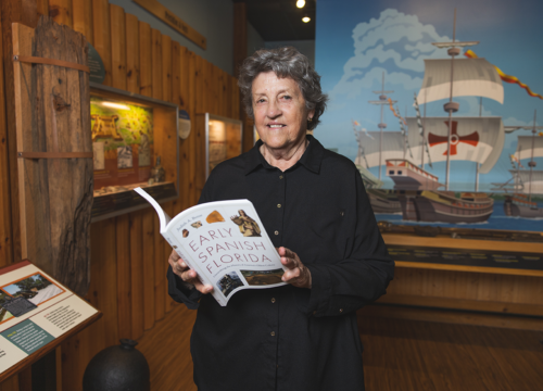 Dr. Judy Bense holds her book, "Early Spanish Florida," inside a museum with a Spanish ship painted on the wall behind her.